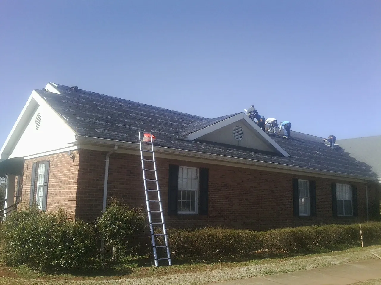 Crew tearing shingles off a brick ranch roof with a ladder set — Emergency Roof Repair work by our crew in Rose