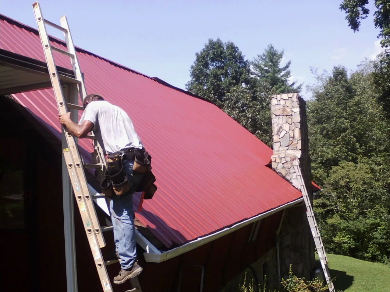 Roofer on a ladder beside a red metal roof and stone chimney — Soffit & Fascia Repair by Roofing Brothers in Rose